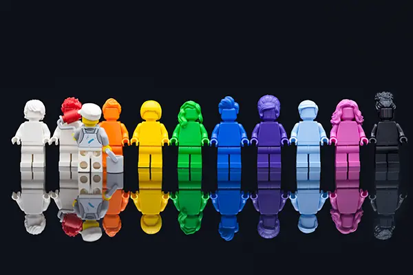 A lineup of LEGO minifigures in solid monochrome colors representing the rainbow Pride flag, standing side by side on a reflective surface.