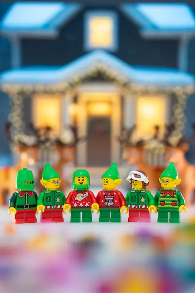 Six LEGO Christmas elf minifigures stand in a row wearing green and red festive outfits, posed in front of a warmly lit snowy workshop with reindeer decorations.