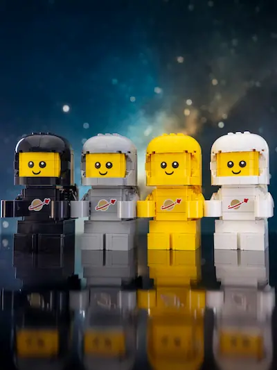 Four custom brick-built LEGO Space Babies in black, gray, yellow, and white spacesuits stand side by side against a starry blue backdrop, with their reflections visible on a glossy surface.