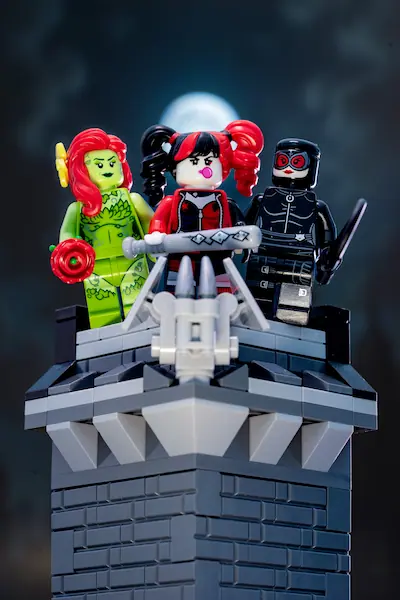 LEGO Poison Ivy, Harley Quinn, and Catwoman pose together on a gothic Gotham rooftop beneath a glowing full moon, with Harley holding a bat and Catwoman carrying a whip.
