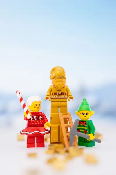 Three Christmas-themed LEGO Build-A-Minifigure characters pose in a snowy scene: a woman holding a candy cane, a gold Santa figure standing on a ladder, and an elf carrying a silver toy sword.