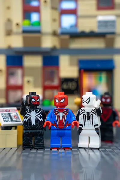 Three custom LEGO Spider-Man minifigures inspired by Insomniac's video games stand together on a city street, wearing the black Symbiote Suit, red-and-blue Advanced Suit, and white Anti-Venom Suit.
