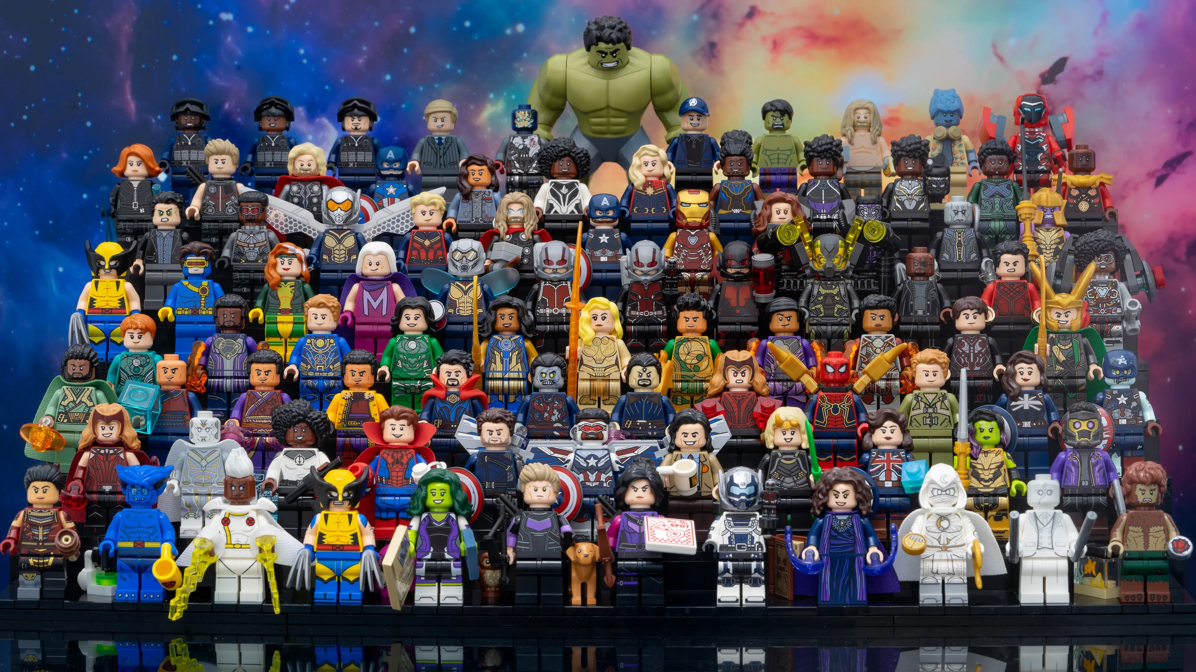 Large group photo of LEGO Marvel minifigures arranged on tiered black steps, featuring characters from across the MCU including Hulk, Spider-Man, Iron Man, Doctor Strange, and Black Panther, set against a colorful cosmic background.
