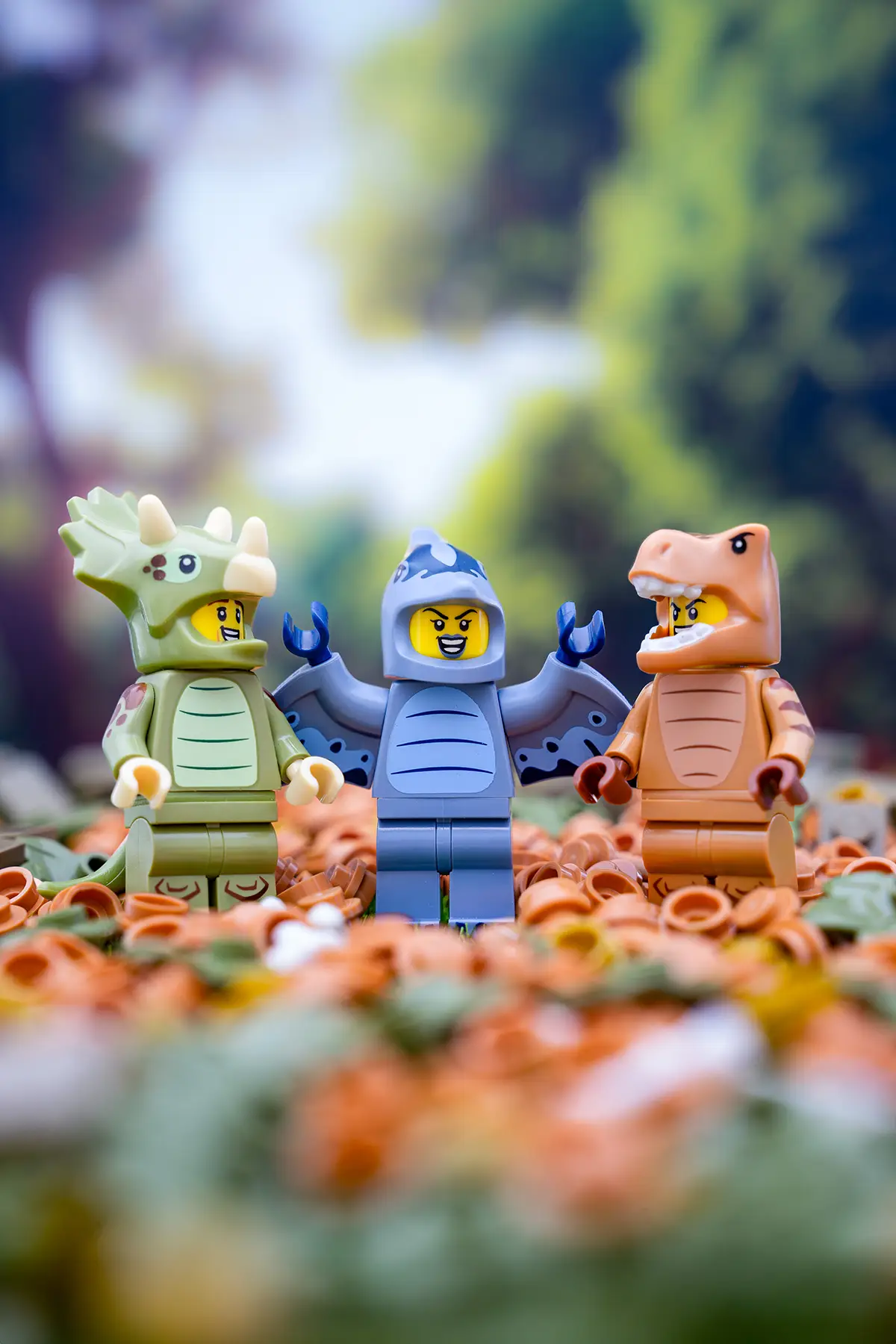 Three LEGO minifigures dressed in dinosaur costumes—triceratops, pteranodon, and T. rex—posing on a bed of LEGO foliage.