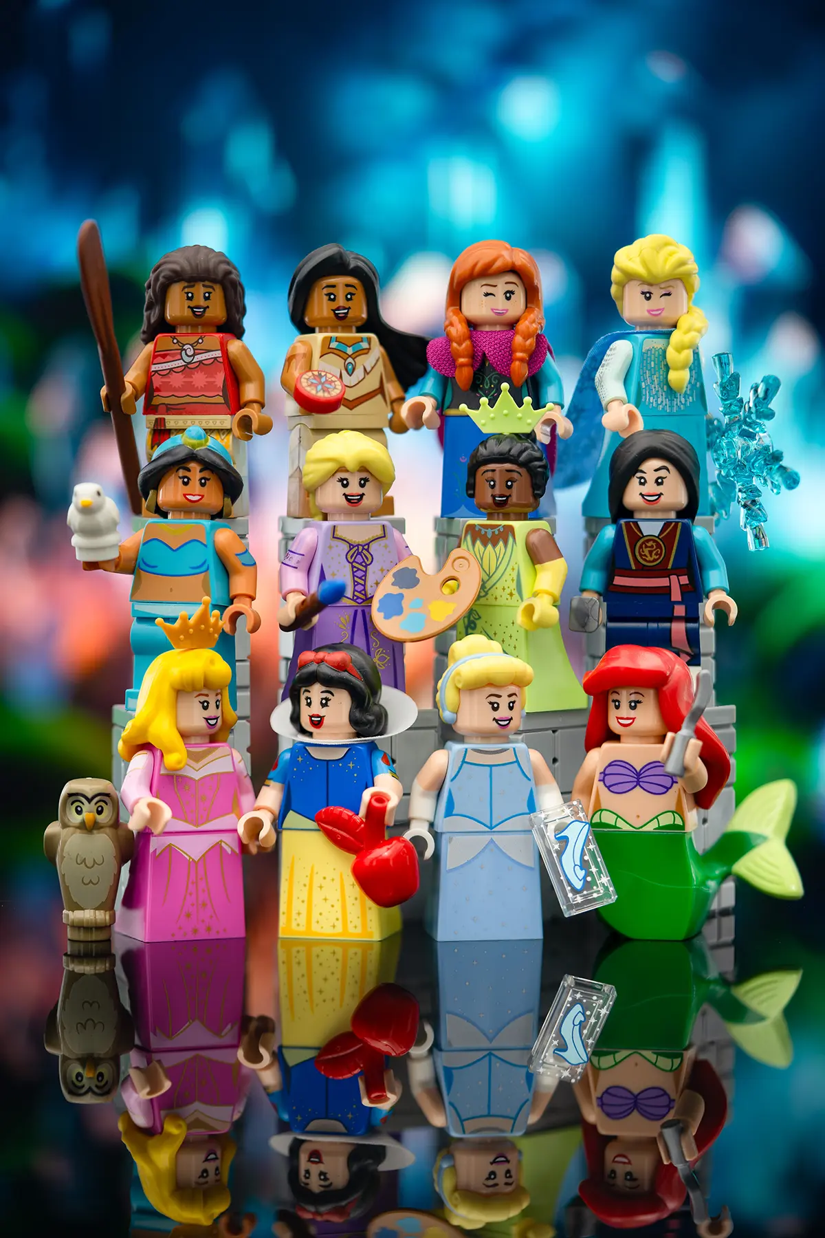 A lineup of LEGO Disney princess minifigures including Snow White, Cinderella, Aurora, Ariel, Jasmine, Belle, Mulan, Tiana, Rapunzel, Moana, Anna, and Elsa, each holding a unique accessory, posed in front of a pastel castle backdrop.