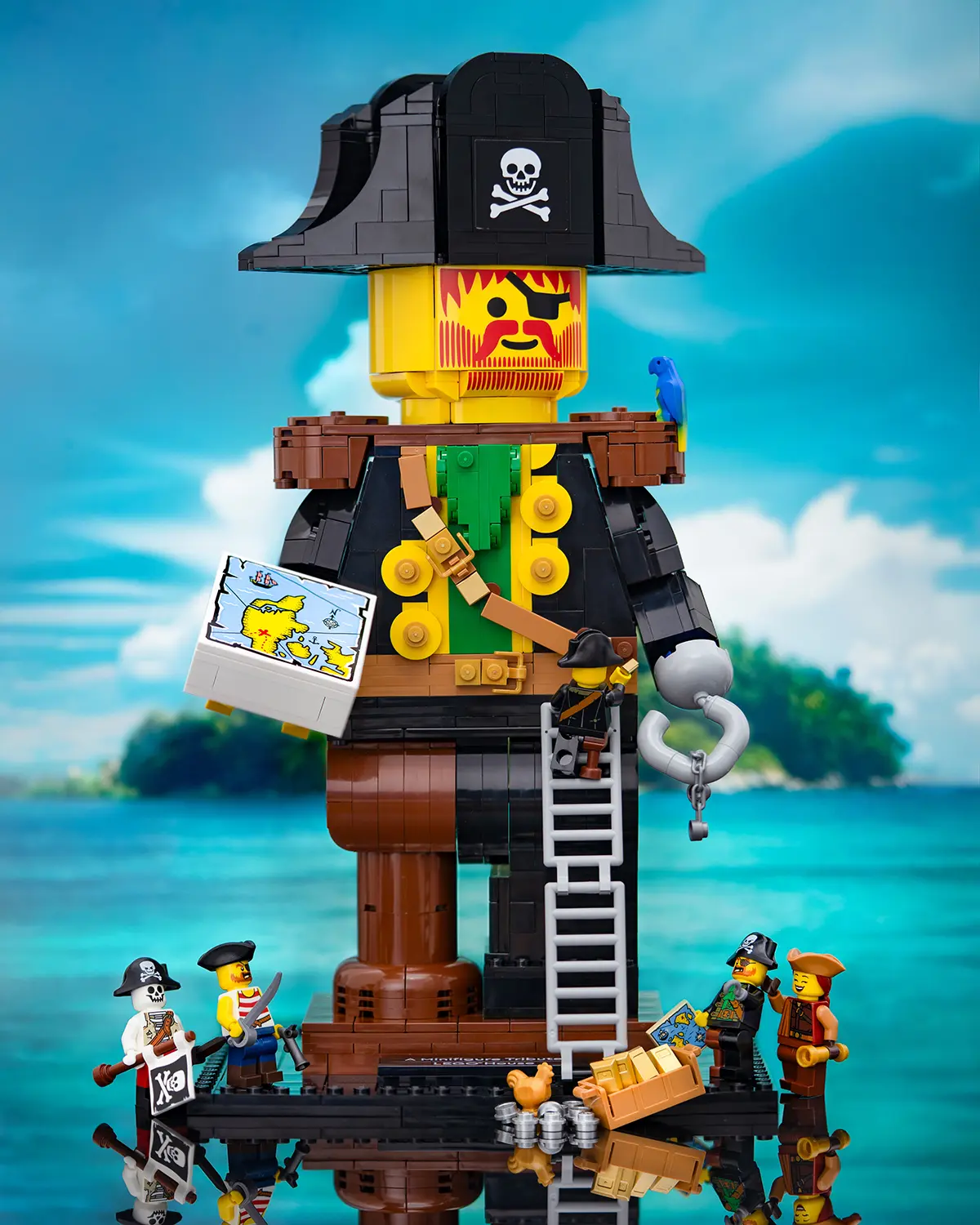 LEGO pirate minifigures build and explore around a giant pirate statue made from bricks, featuring an eye patch, hook hand, and a treasure map. The scene is set against a bright tropical ocean backdrop, with palm trees and an island in the distance.