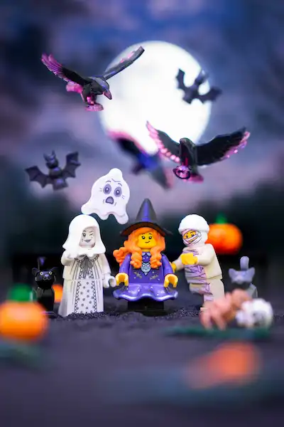 LEGO Halloween scene with a witch minifigure, a white ghost figure, and a mummy under a bright full moon with bats flying overhead.