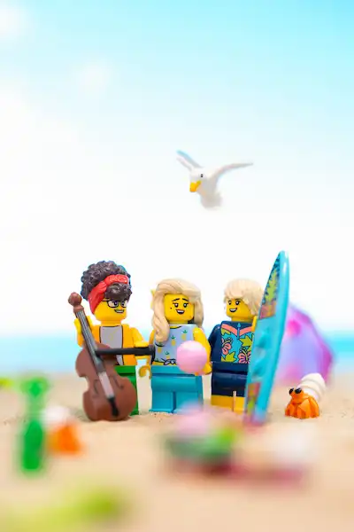 Three LEGO minifigures on a sandy beach scene with a guitar, a surfboard, and a small crab, with a seagull flying above.