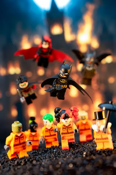 LEGO Batman and members of the Bat-Family leap into action above escaped Arkham inmates, including Harley Quinn, the Joker, Scarecrow, and the Penguin, as flames engulf the asylum in the background.