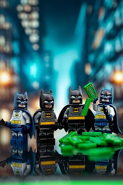 A group of LEGO Batman minifigures in various costumes stand proudly with a pile of LEGO cash in a city backdrop.
