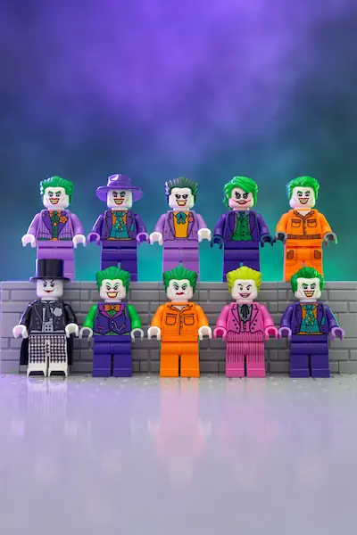 Ten LEGO Joker minifigure variants are arranged in two rows against a gray brick wall, featuring different purple, orange, pink, and black outfits with green hair and distinctive smiling faces.