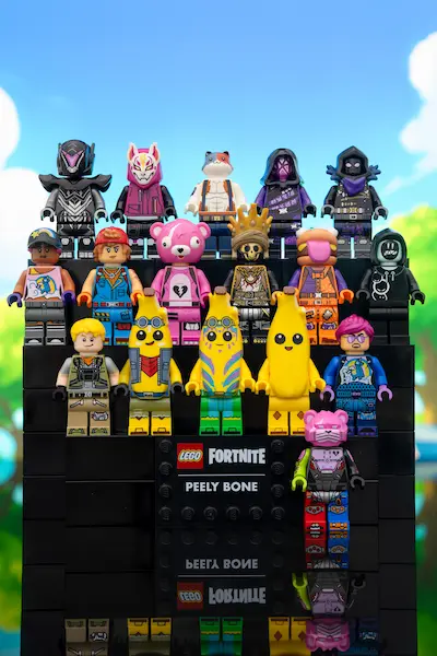 Group photo of LEGO Fortnite minifigures arranged on black display steps, with Peely variants and multiple characters posed in a lineup against a bright sky background.