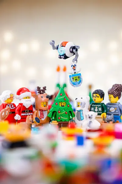 LEGO Advent Calendar 2025 scene with festive minifigures gathered around a Christmas tree costume figure, including Santa, a reindeer suit character, a polar bear costume, and holiday sweater minifigs.
