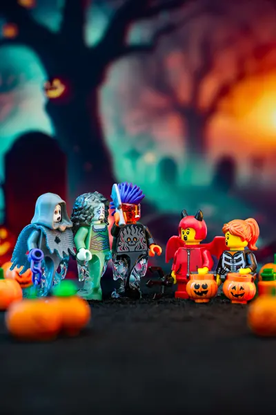 LEGO minifigures in Halloween costumes including a ghost, zombie, devil, and skeleton gather with pumpkin buckets in a spooky graveyard setting.