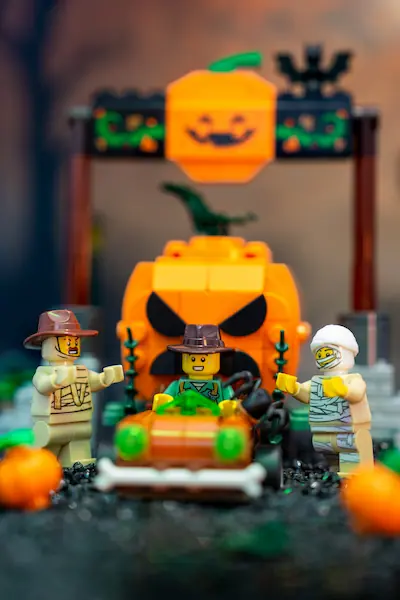 LEGO Halloween scene featuring two mummy minifigures and an explorer surrounding a pumpkin vehicle, with a giant angry jack-o'-lantern build and glowing Halloween decorations in the background.