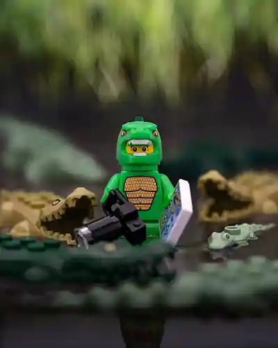 	LEGO minifigure in a green crocodile costume standing in a swampy setting surrounded by crocodile figures, holding a map and camera.