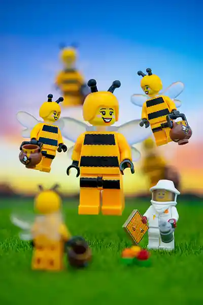 LEGO bee-costumed minifigures flying joyfully around a beekeeper holding a honeycomb tile on a grassy field at sunset.