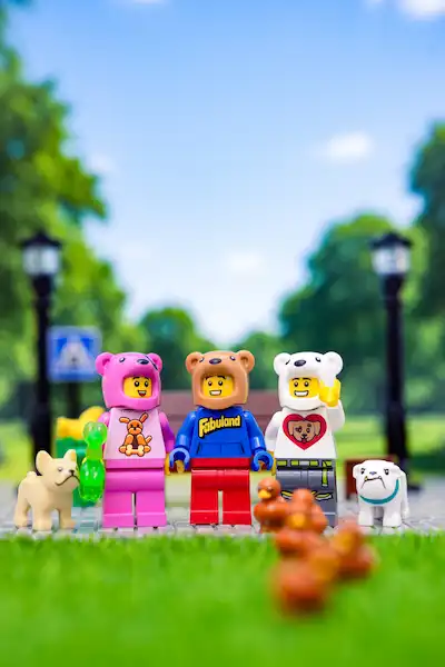 Three LEGO minifigures in bear hoodies stand on a sidewalk in a park scene, with two small bulldog figures beside them and a blurred squirrel in the foreground.