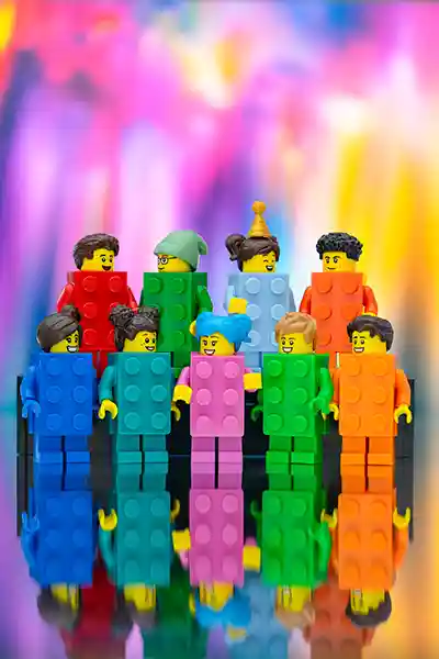 A lineup of LEGO minifigures dressed in colorful brick suits—red, blue, yellow, and green—stand together in front of a vibrant cityscape, celebrating LEGO creativity and fun.