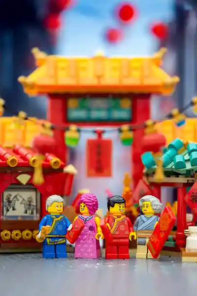 LEGO minifigures dressed in traditional Chinese attire exchanging red envelopes in front of a festive Lunar New Year street scene with lanterns and a red gate.