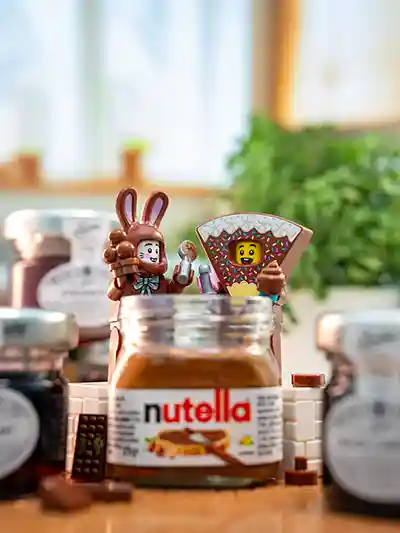 LEGO minifigures enjoy a breakfast scene with a giant brick-built Nutella jar, surrounded by toast, croissants, and fruit on a sunny kitchen table.