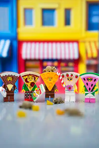 LEGO minifigures dressed in food-themed costumes including pizza, watermelon, cheese, and chocolate, standing on a street with colorful storefronts behind them.