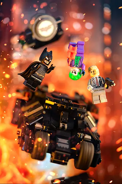 LEGO Batman jumps from the Tumbler amidst fiery explosions, confronting Joker and Two-Face.