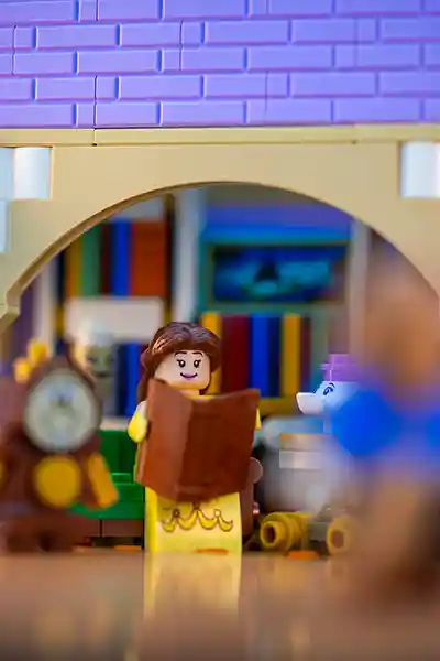 LEGO Belle stands in a grand library, surrounded by towering bookshelves and enchanted objects, holding a book and smiling warmly.