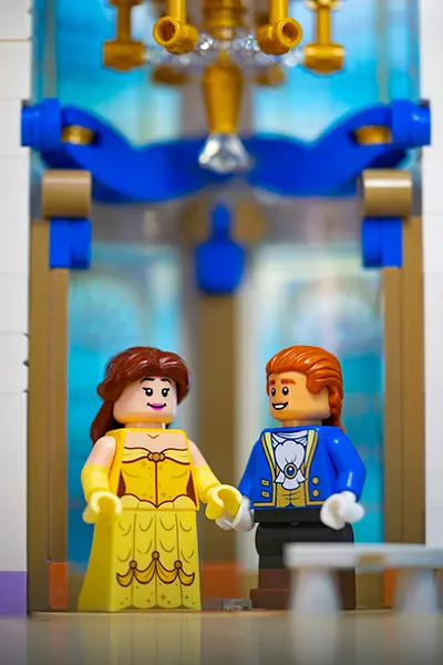 LEGO minifigures of Belle in her yellow gown and Prince Adam in a blue suit, standing beneath a golden chandelier in a castle ballroom.