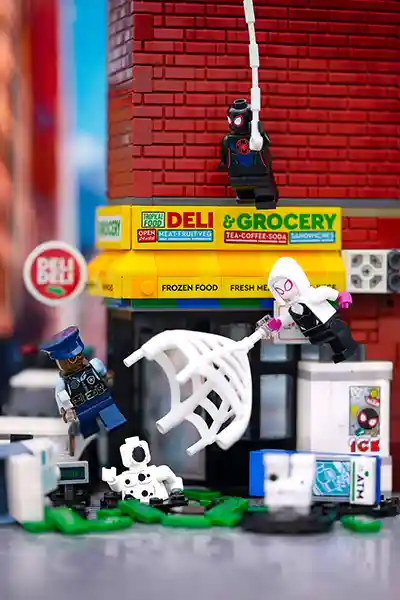 LEGO Miles Morales swings above a city deli while Spider-Gwen webs a villain near a fallen police officer and ATM debris.