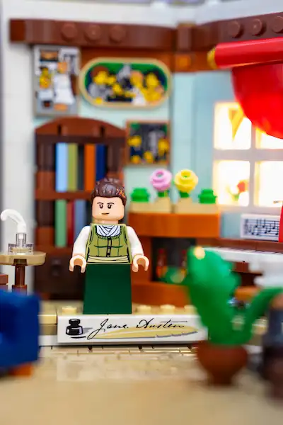 LEGO Jane Austen minifigure stands in a detailed study with books, flowers, and a piano.