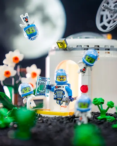 A LEGO astronaut at a moonbase is surrounded by playful green aliens, with the moon glowing in the background.