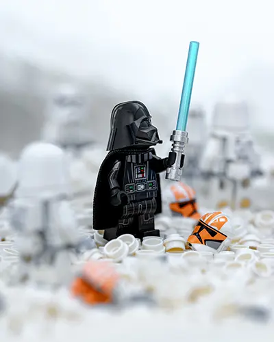 LEGO Darth Vader stands solemnly in the snow among fallen clone helmets, holding a blue lightsaber—recreating the Clone Wars finale.